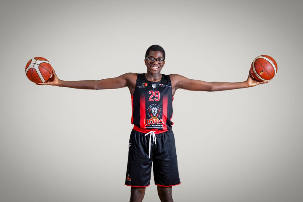 Basketball player in dark jersey poses with basketballs outstretched against grey background.