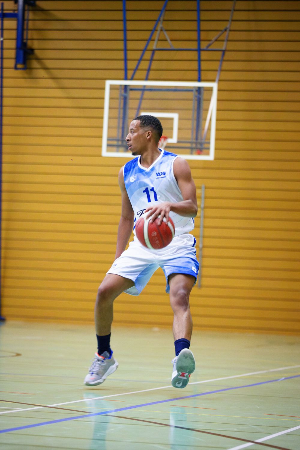Basketball player in white jersey number 11 dribbles on indoor court against yellow wall backdrop.