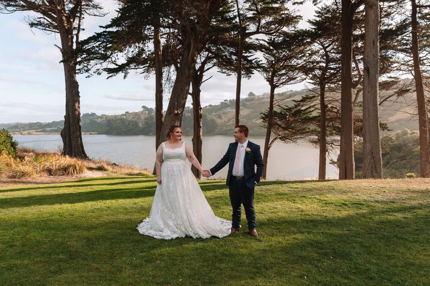 Wedding couple poses on grassy lawn with tall pine trees and water view in background.