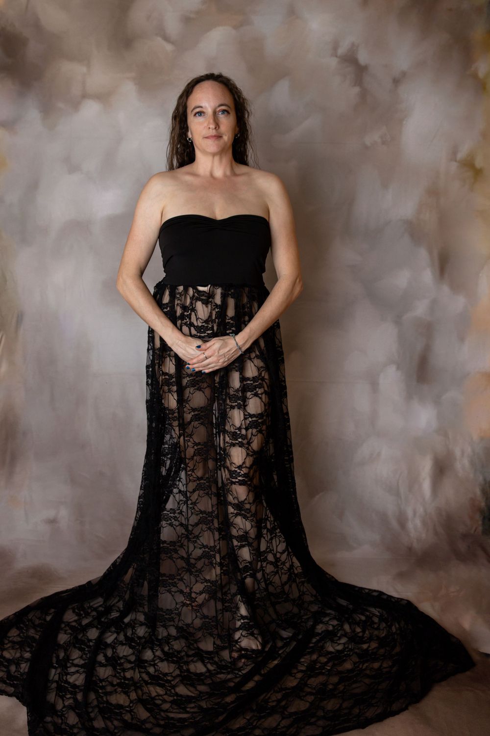 Woman in elegant black strapless lace gown with floral pattern posing against neutral painted backdrop during maternity photography session.