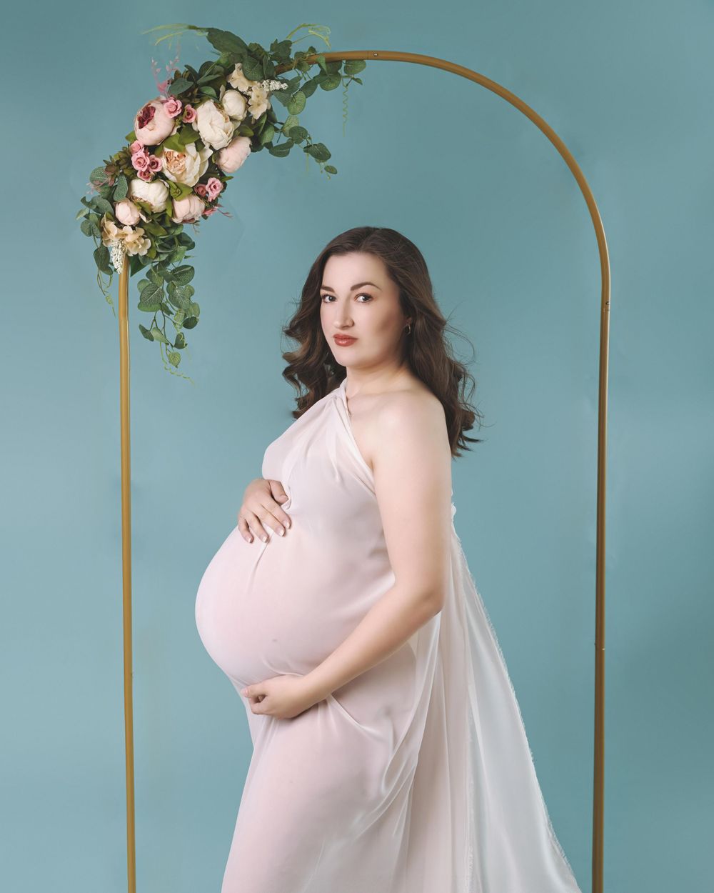 A maternity photo showing an expectant mother in a sheer white dress posing next to a gold arch with floral decorations.