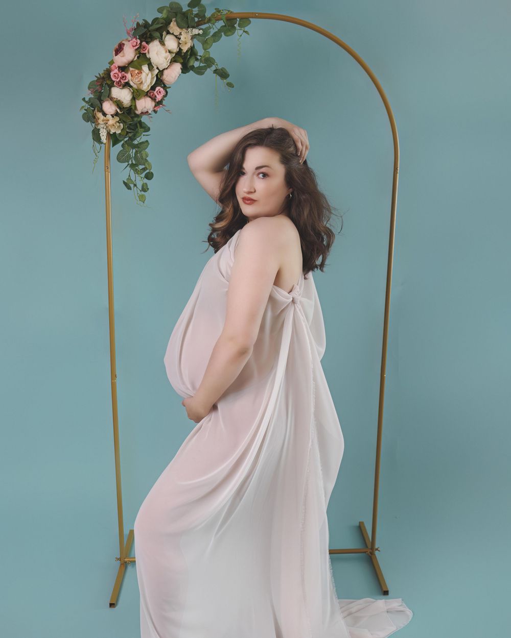 Ethereal maternity portrait with flowing white dress against blue backdrop, featuring gold arch decorated with pink flowers.