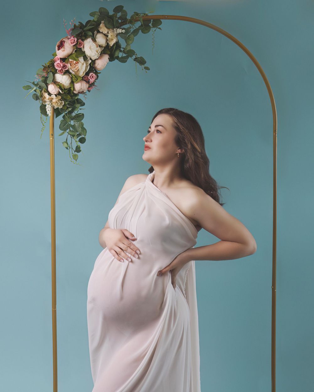 Ethereal maternity photo featuring flowing white gown against turquoise wall with golden arch and floral arrangement.