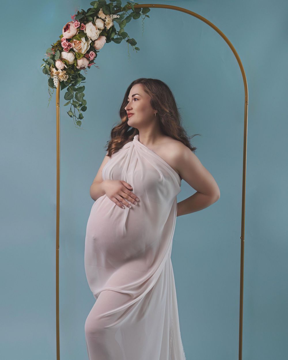 A serene maternity photo with a flowing white dress against a blue wall backdrop and gold arch decorated with pink flowers.