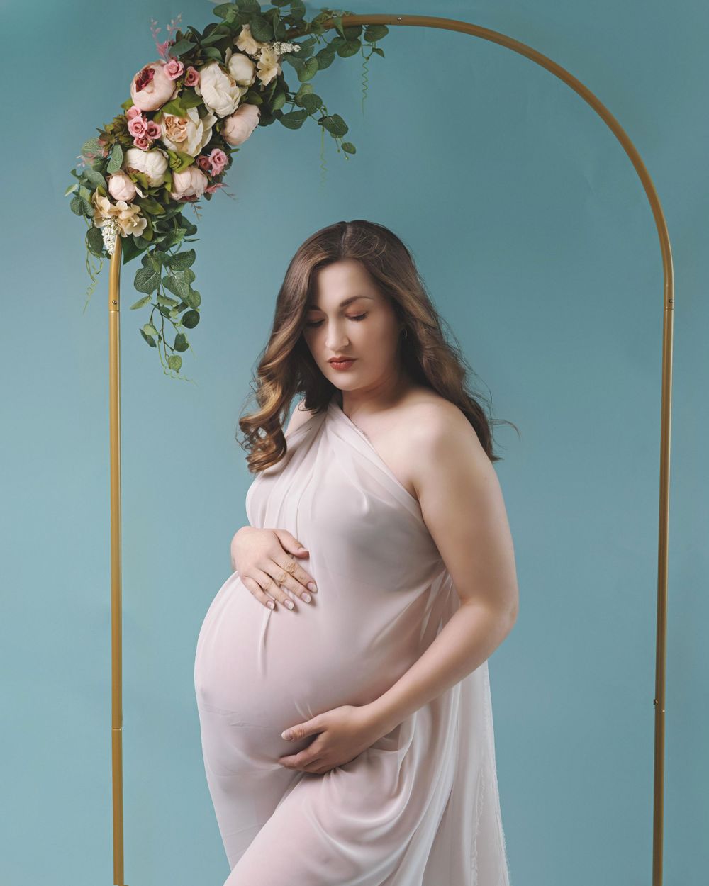 A dreamy maternity portrait against a turquoise backdrop features a pink dress and gold arch adorned with roses and greenery.