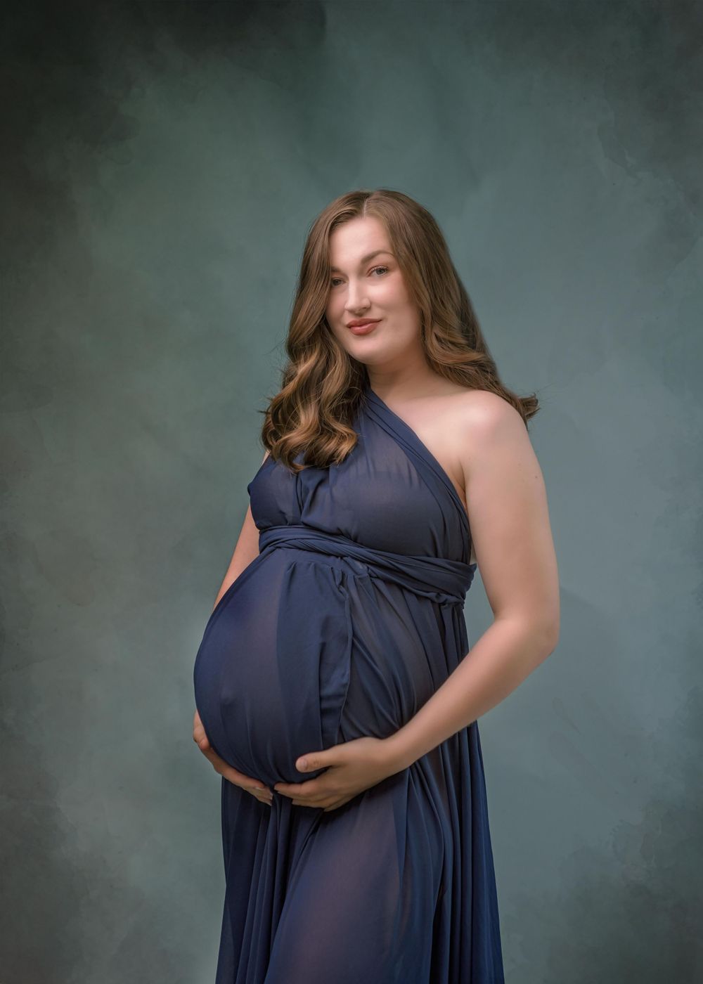 Studio maternity portrait of an expectant mother in a flowing navy blue dress against a dark teal backdrop.