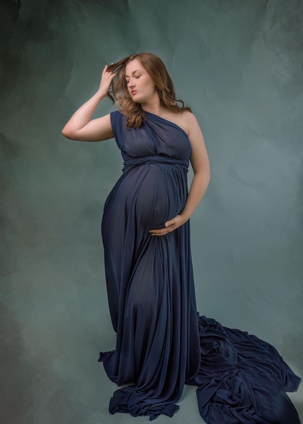 A pregnant woman poses in an elegant navy blue one-shoulder maternity gown against a dark moody background.