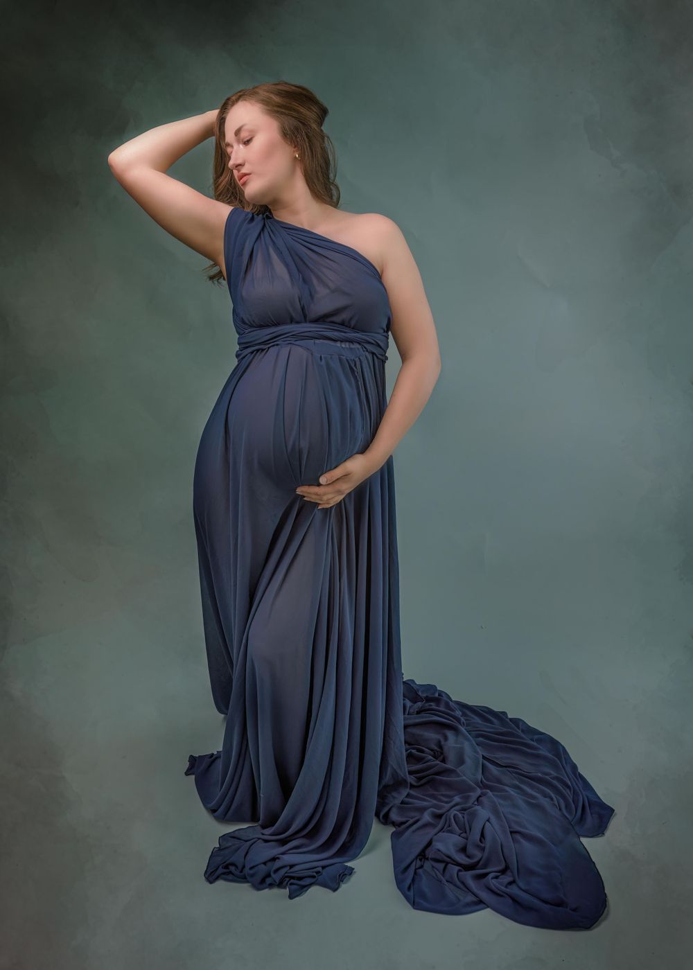 A one-shoulder navy blue maternity gown flows elegantly against a moody dark backdrop in an artistic portrait.