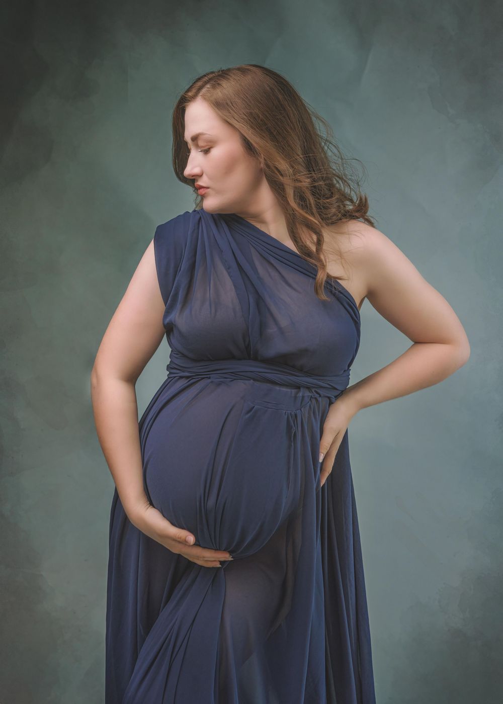 Side profile of someone wearing a flowing navy gown in a maternity photo with a moody gray studio backdrop.