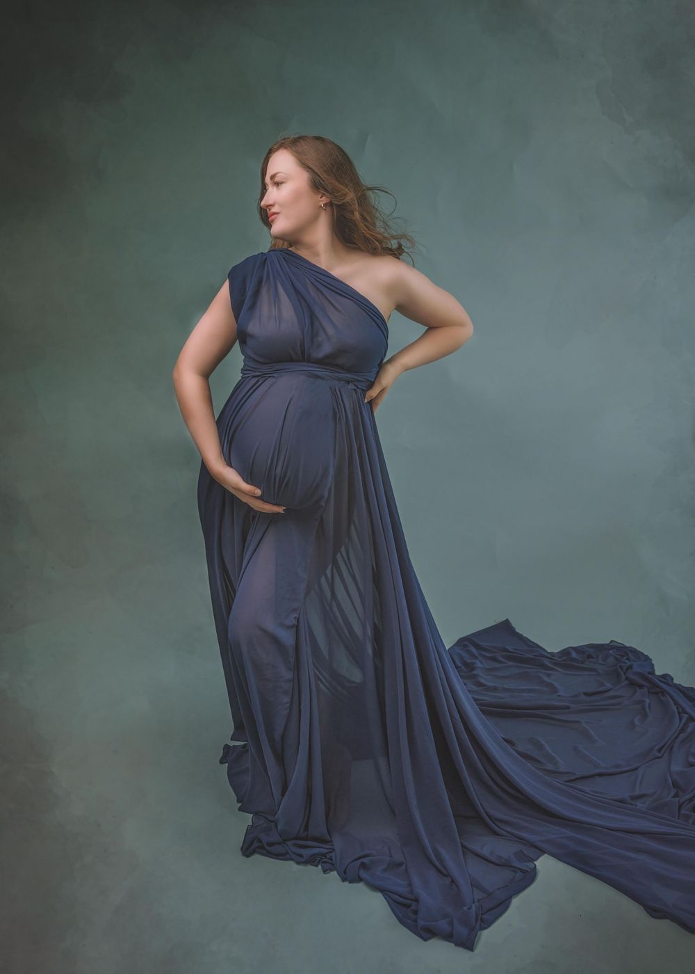 Elegant maternity portrait in navy blue one-shoulder gown with flowing train against moody green background.