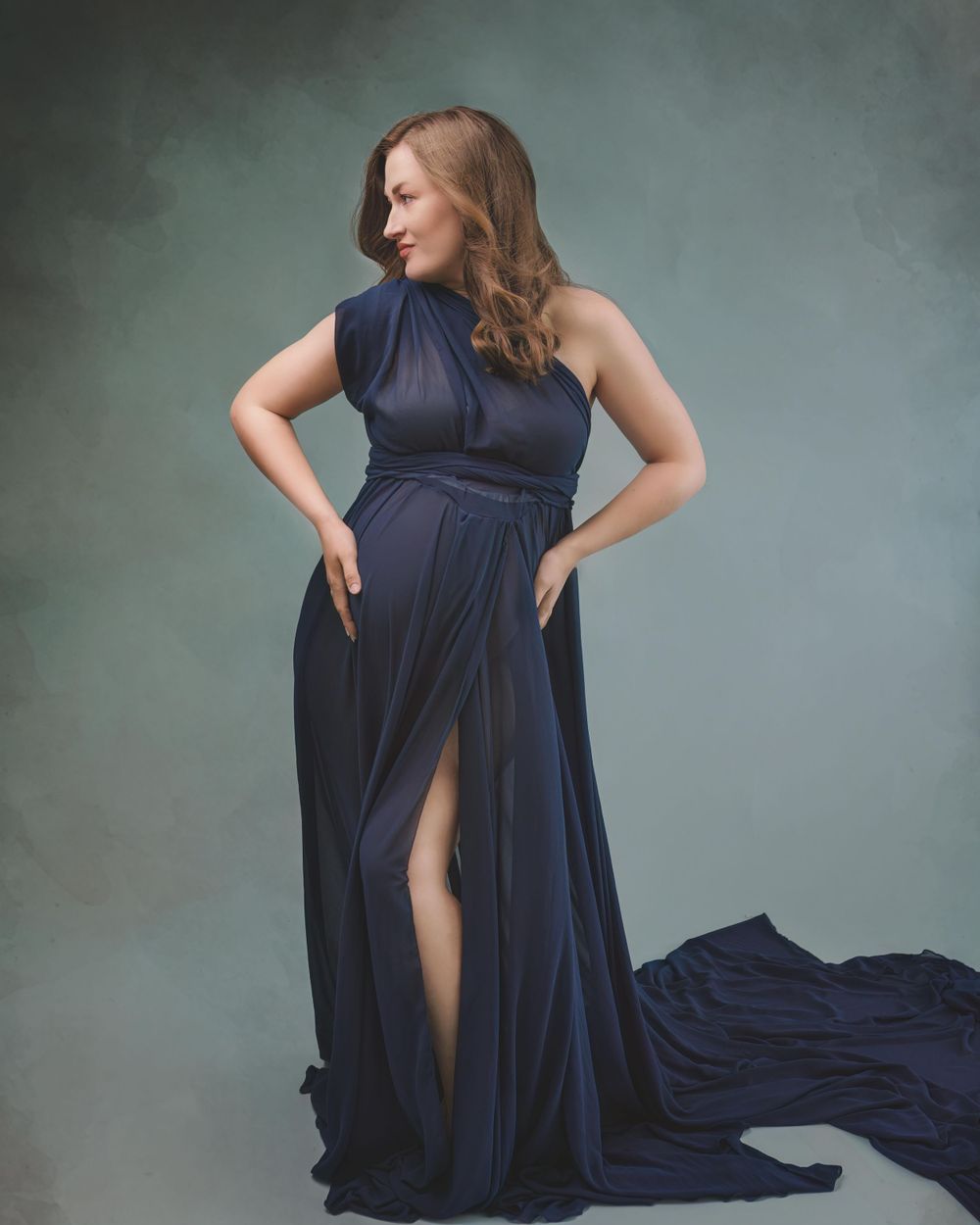 A glamorous navy blue evening gown with a thigh-high slit flows elegantly in a moody studio portrait setting.