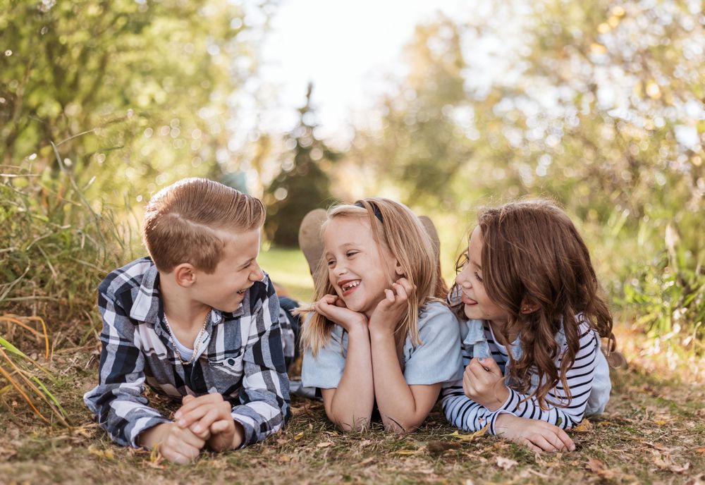 Three children lay on the ground outdoors in casual clothing smiling and playing together in autumn sunlight.
