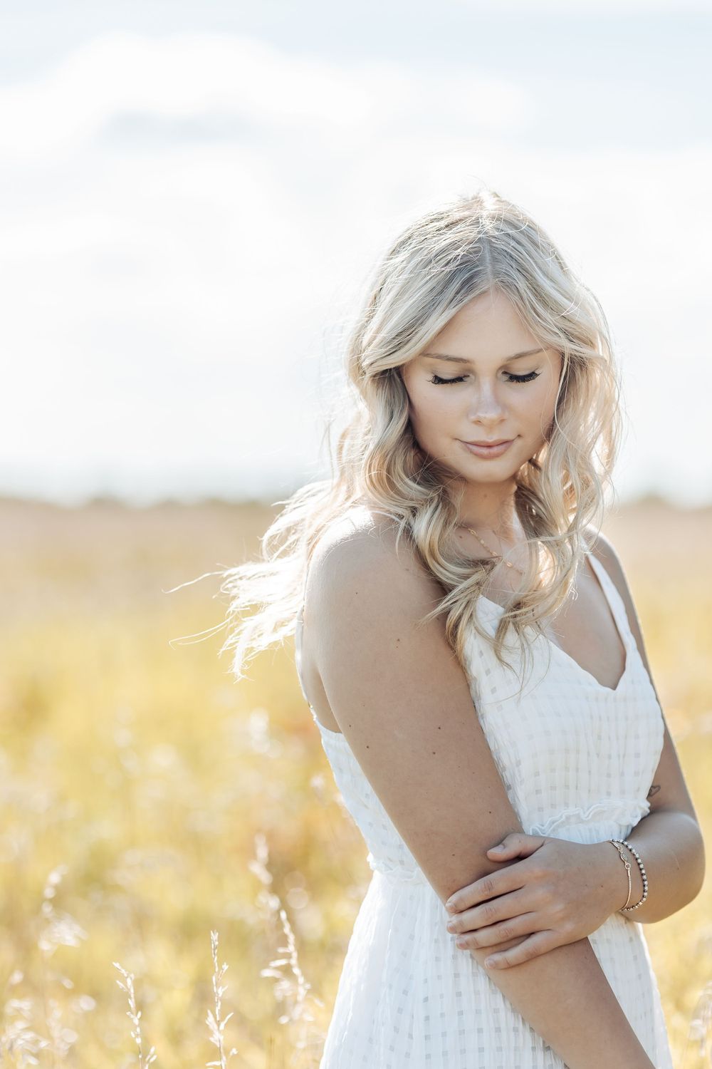 Ethereal portrait of woman in white dress standing in sunlit golden field with soft wind-blown hair.