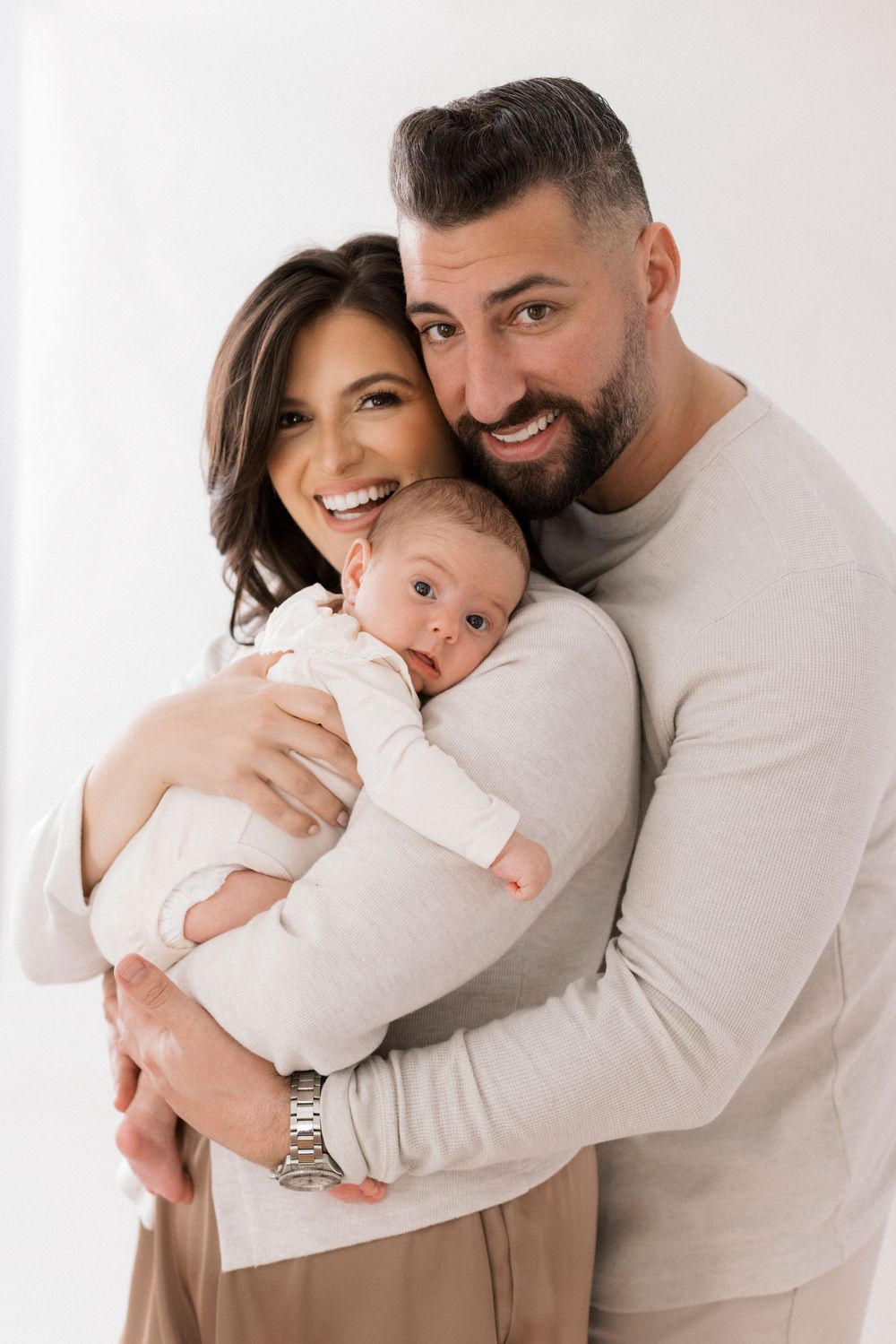 A loving family portrait with parents embracing their baby in neutral-colored clothing against a light background.