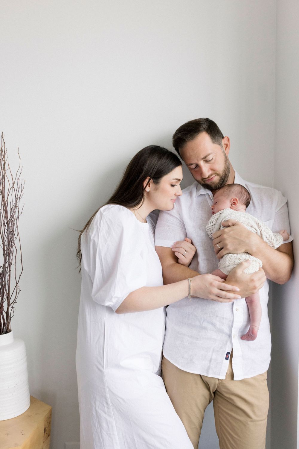 A tender family moment as parents in white clothing cuddle with their newborn baby against a light colored wall.