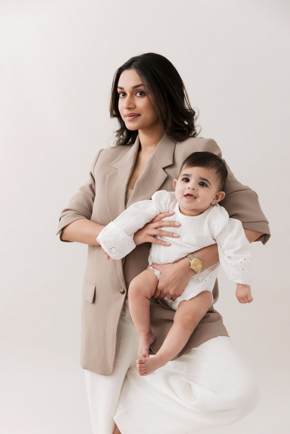 Elegant woman in beige suit holding a smiling baby in white outfit against a light background.