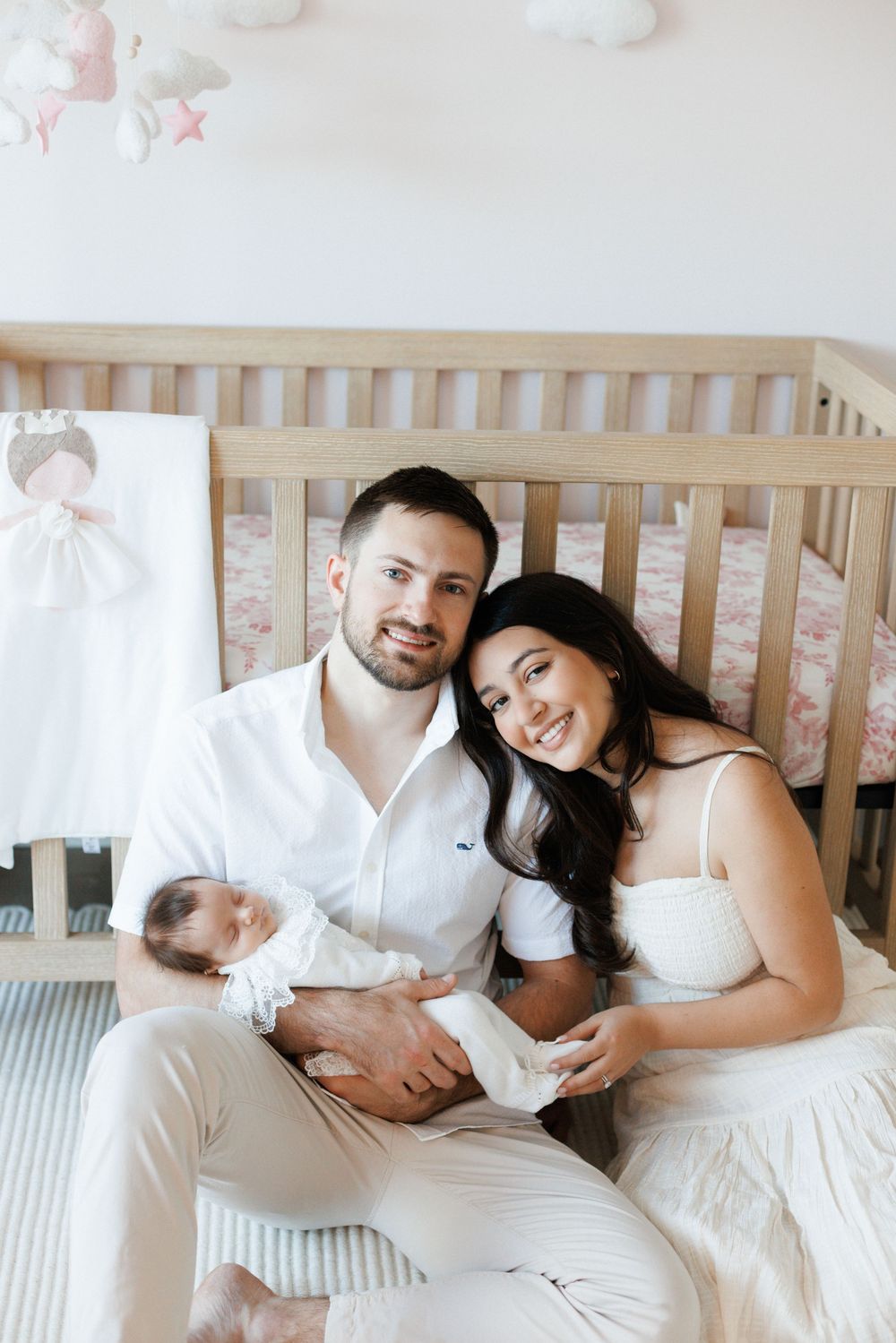 Happy couple sitting in nursery holding their newborn baby, dressed in white, smiling in front of a wooden crib.