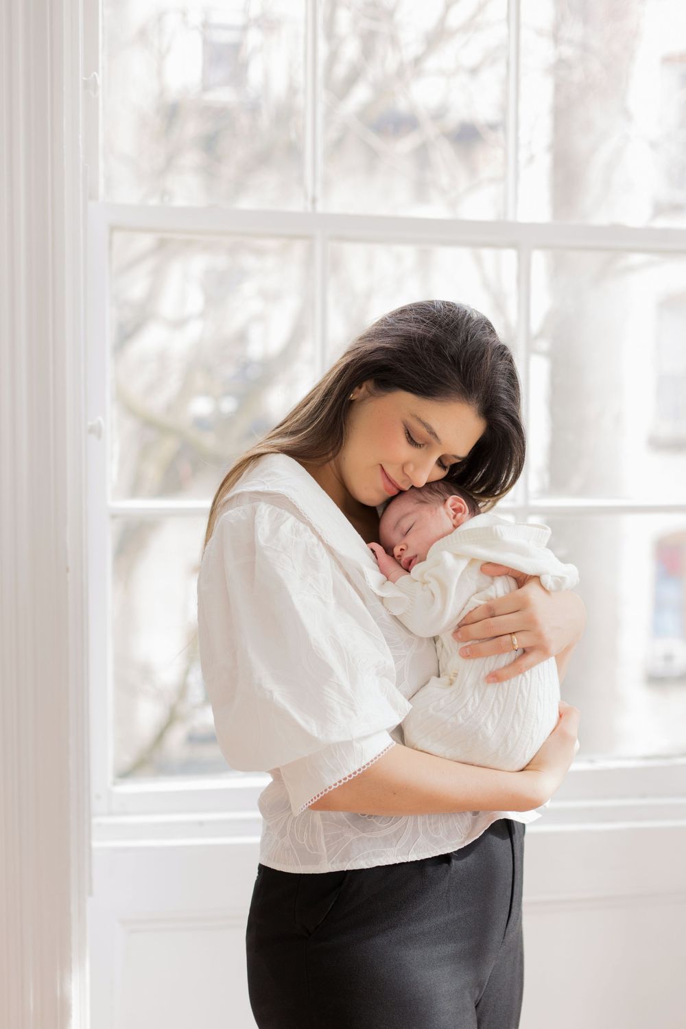 A mother tenderly holds her sleeping newborn baby wrapped in white near a bright window.
