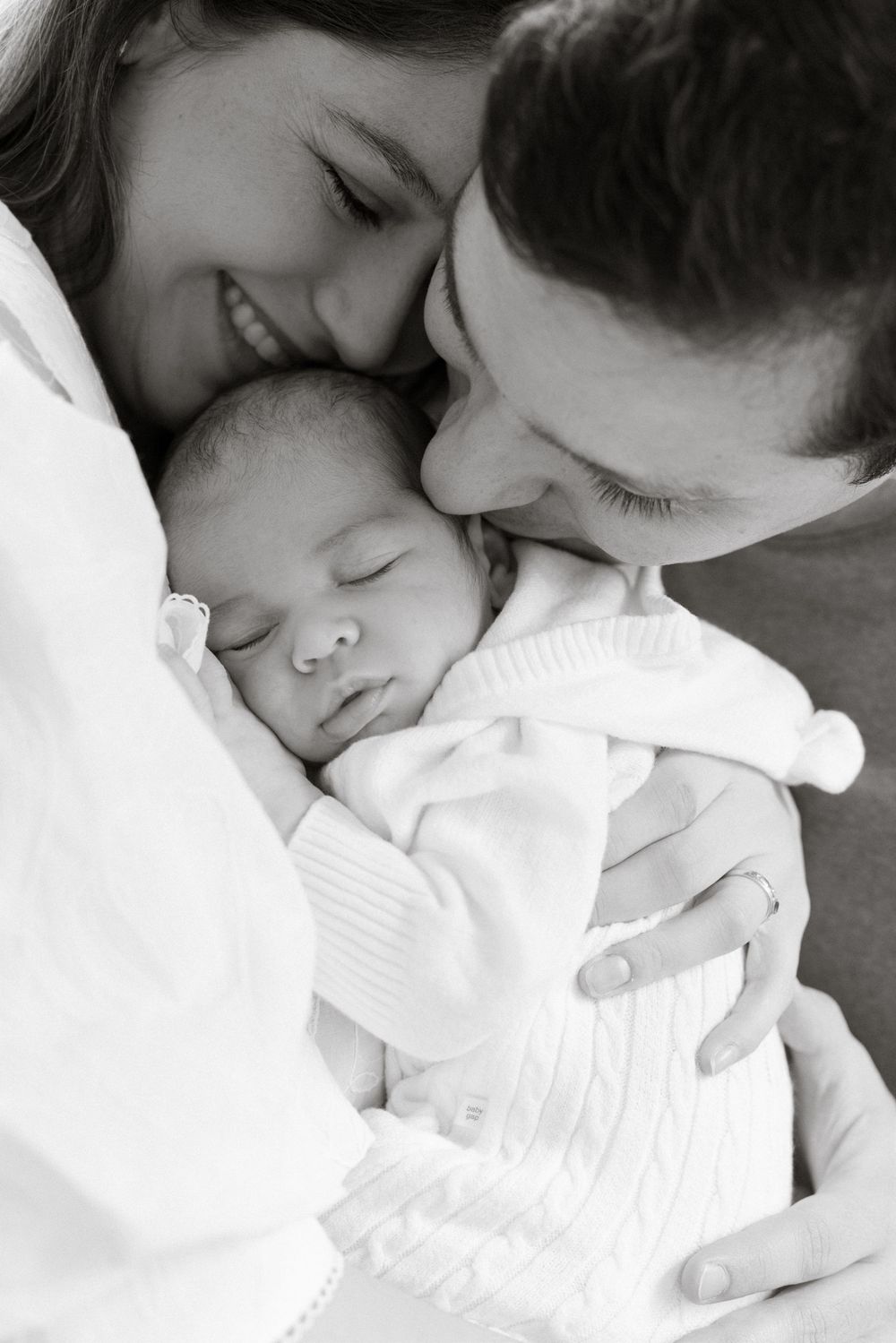 Black and white photo of smiling parents tenderly kissing their sleeping newborn baby wrapped in white.