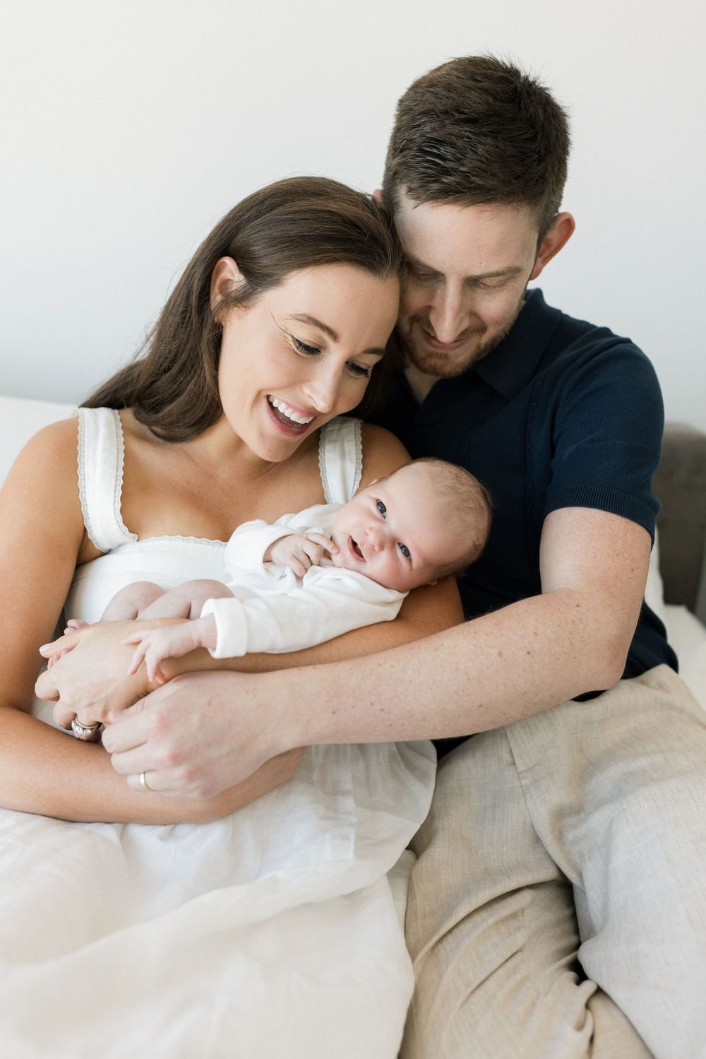 Happy new parents smiling and cuddling their newborn baby dressed in white while sitting together on a bed.