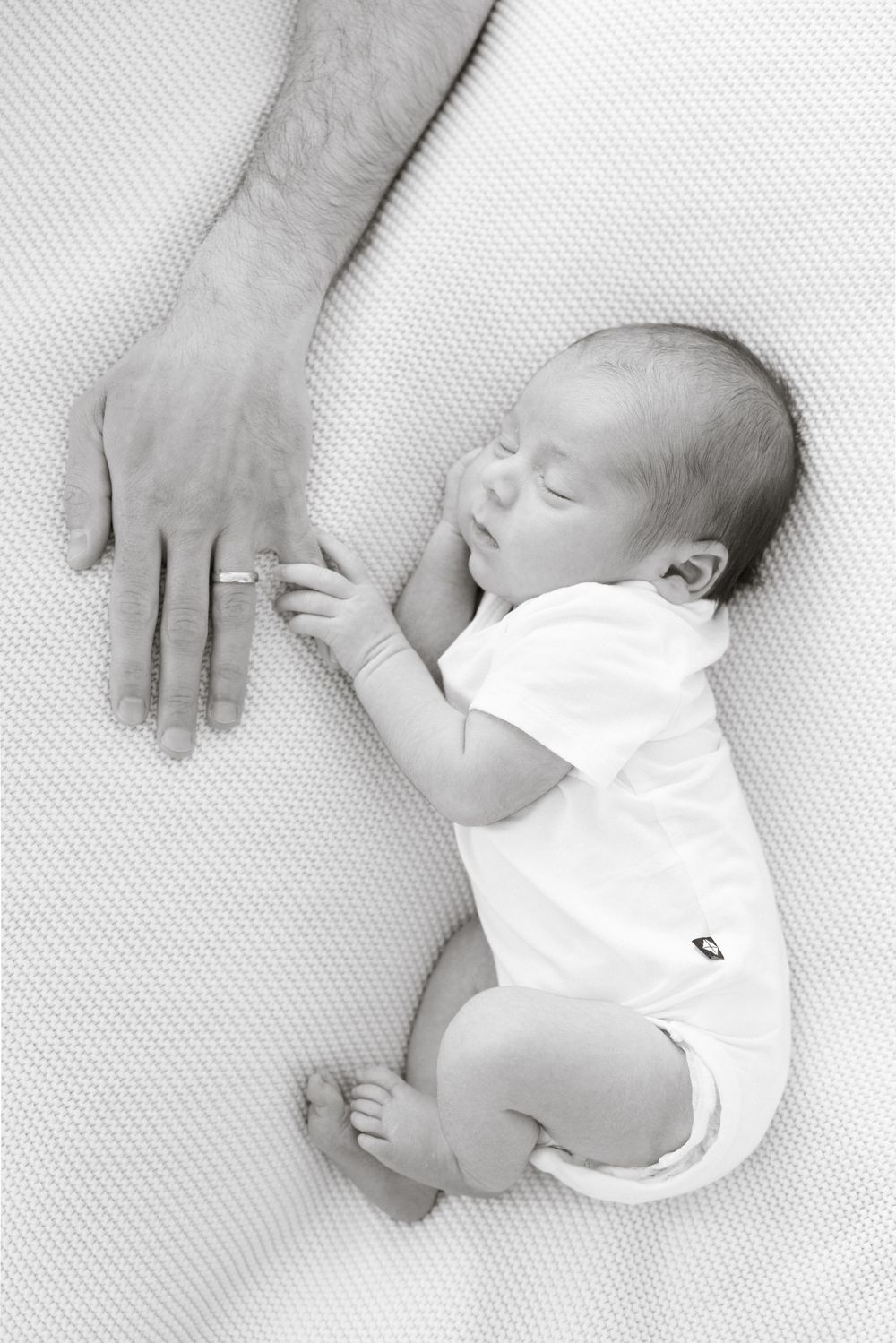 Black and white photo of a sleeping newborn baby holding an adult's finger on a white blanket.