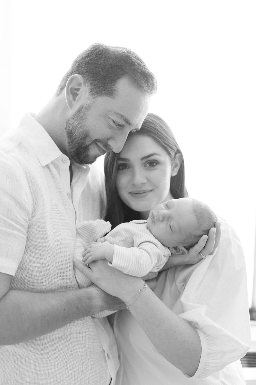 A tender black and white portrait of parents lovingly cradling their newborn baby in matching white outfits.