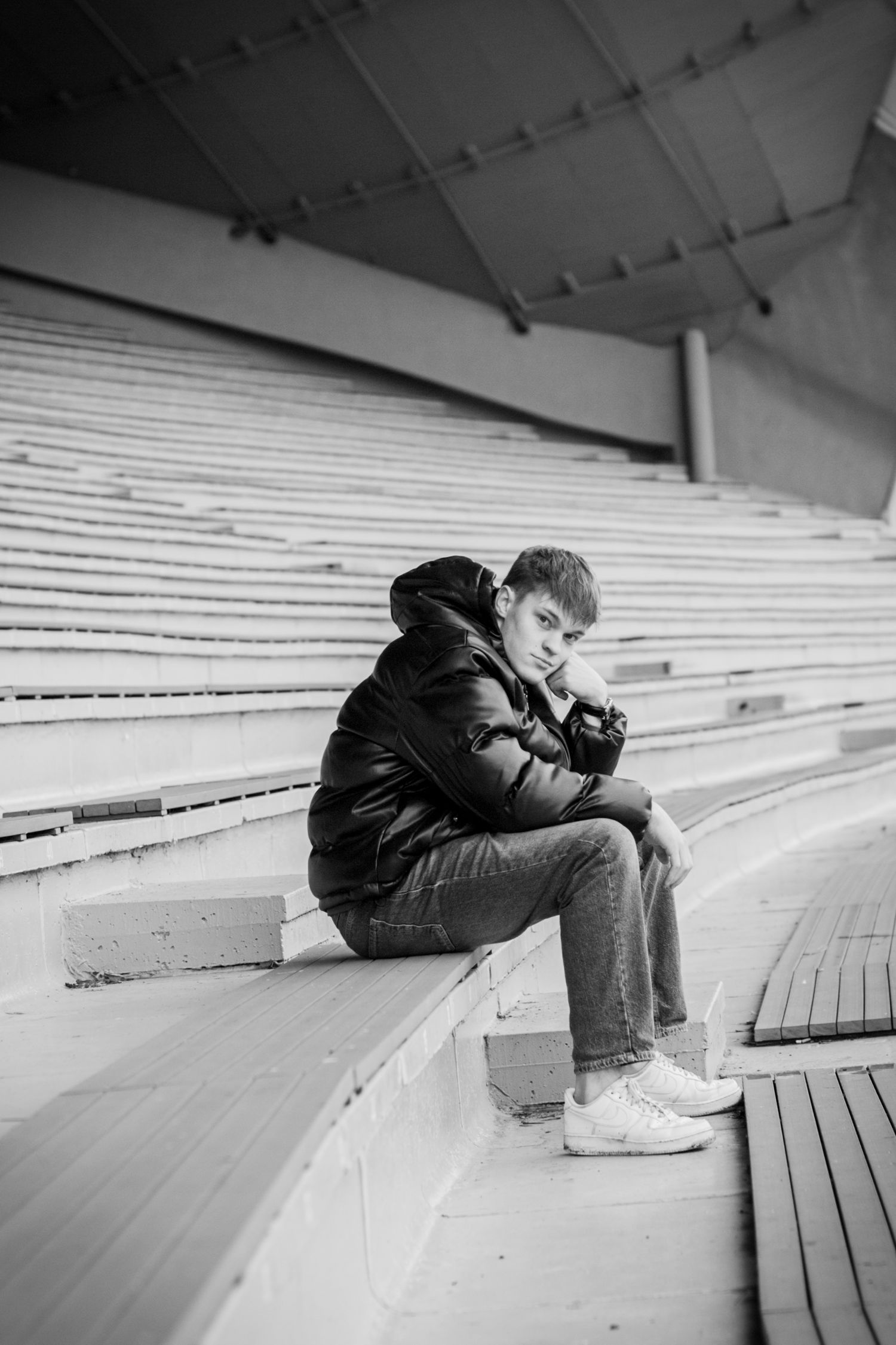Person in black puffer jacket sits alone on wooden stadium bleachers in black and white photo.