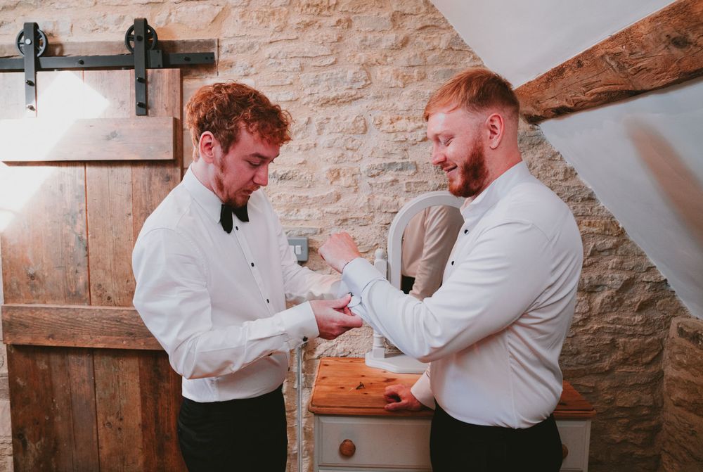 Two men in formal attire helping each other get ready in rustic stone-walled room.