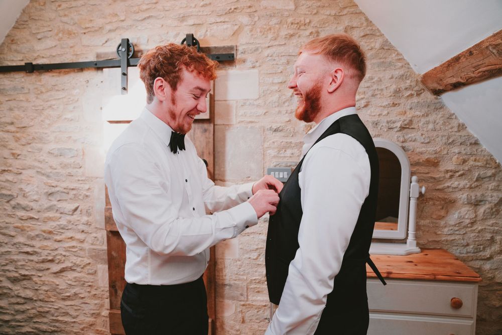 Best man assists with groom's preparation, adjusting tie in stone-walled dressing room.