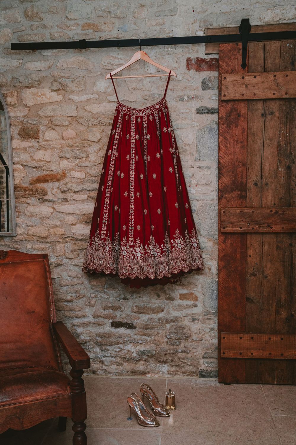 Traditional red and gold Indian lehenga dress hanging against stone wall with shoes below.