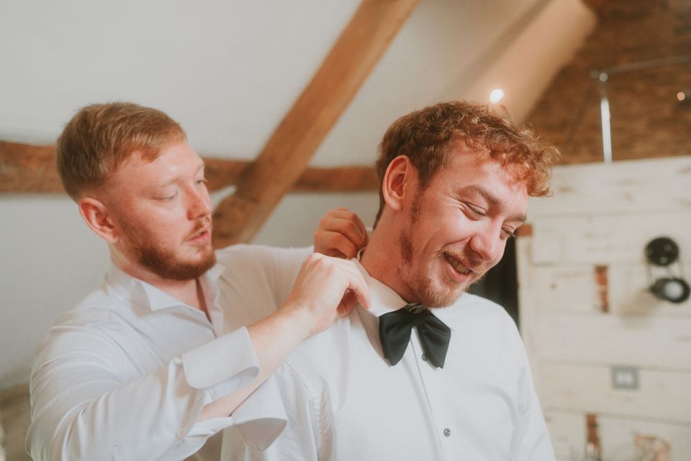 Wedding preparation scene with bow tie adjustment in rustic setting.