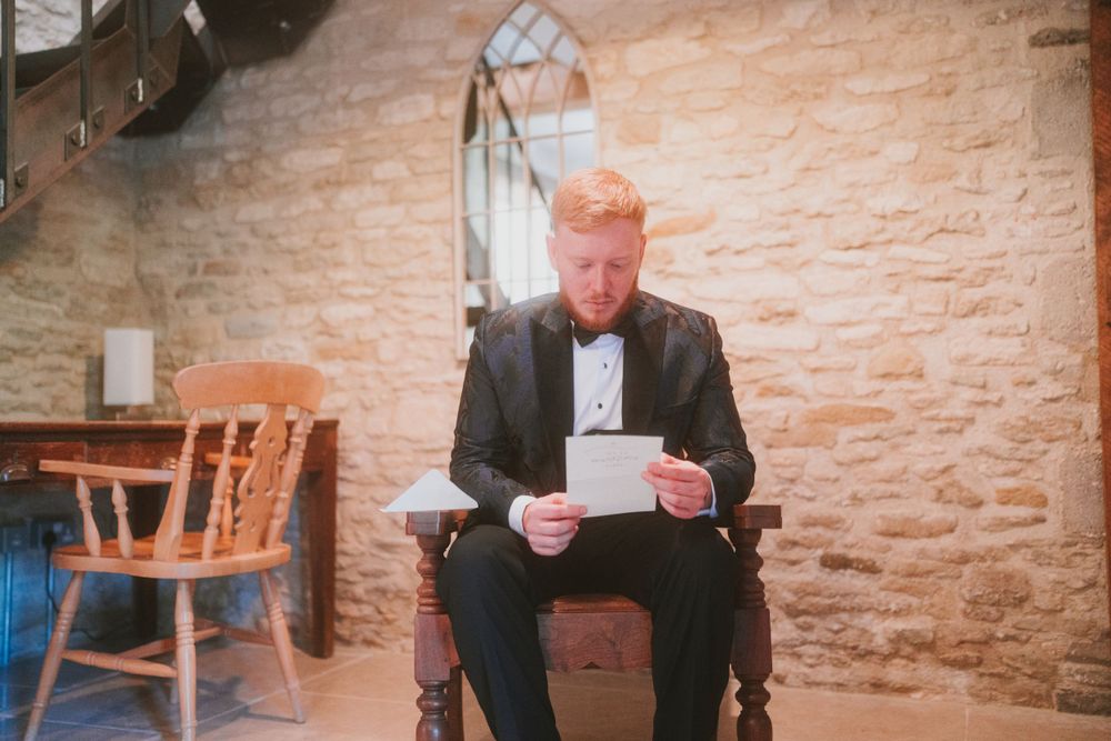 A person sitting in a wooden chair reading from papers against a stone wall.