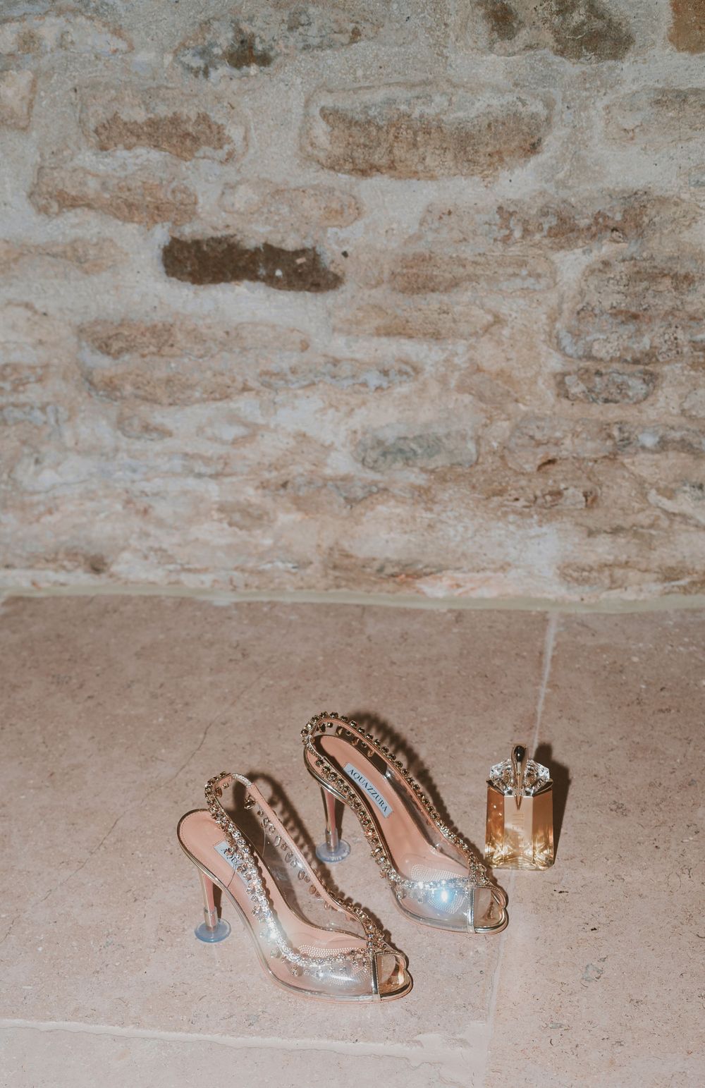 Elegant crystal-embellished bridal shoes and perfume bottle against stone wall background.