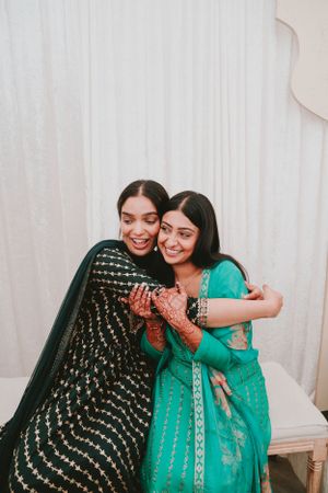 Two women in traditional Indian outfits sharing a joyful embrace against white backdrop.