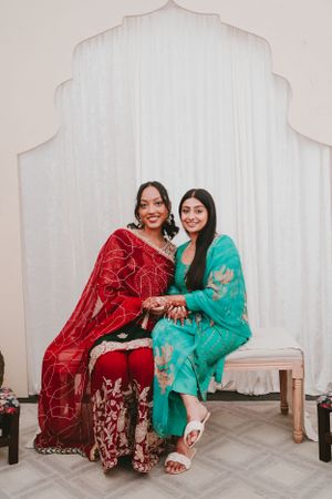 Two women in traditional Indian attire, red and turquoise, seated on bench against white backdrop.