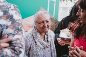 Elderly woman in gray shawl smiling while being served treats at a celebration gathering.