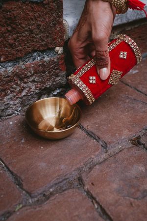 Hand pouring liquid from decorative red vessel into brass bowl on brick surface.