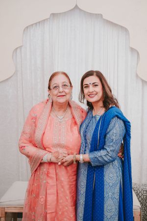 Two people in traditional formal attire pose together against white backdrop with decorative arch design.