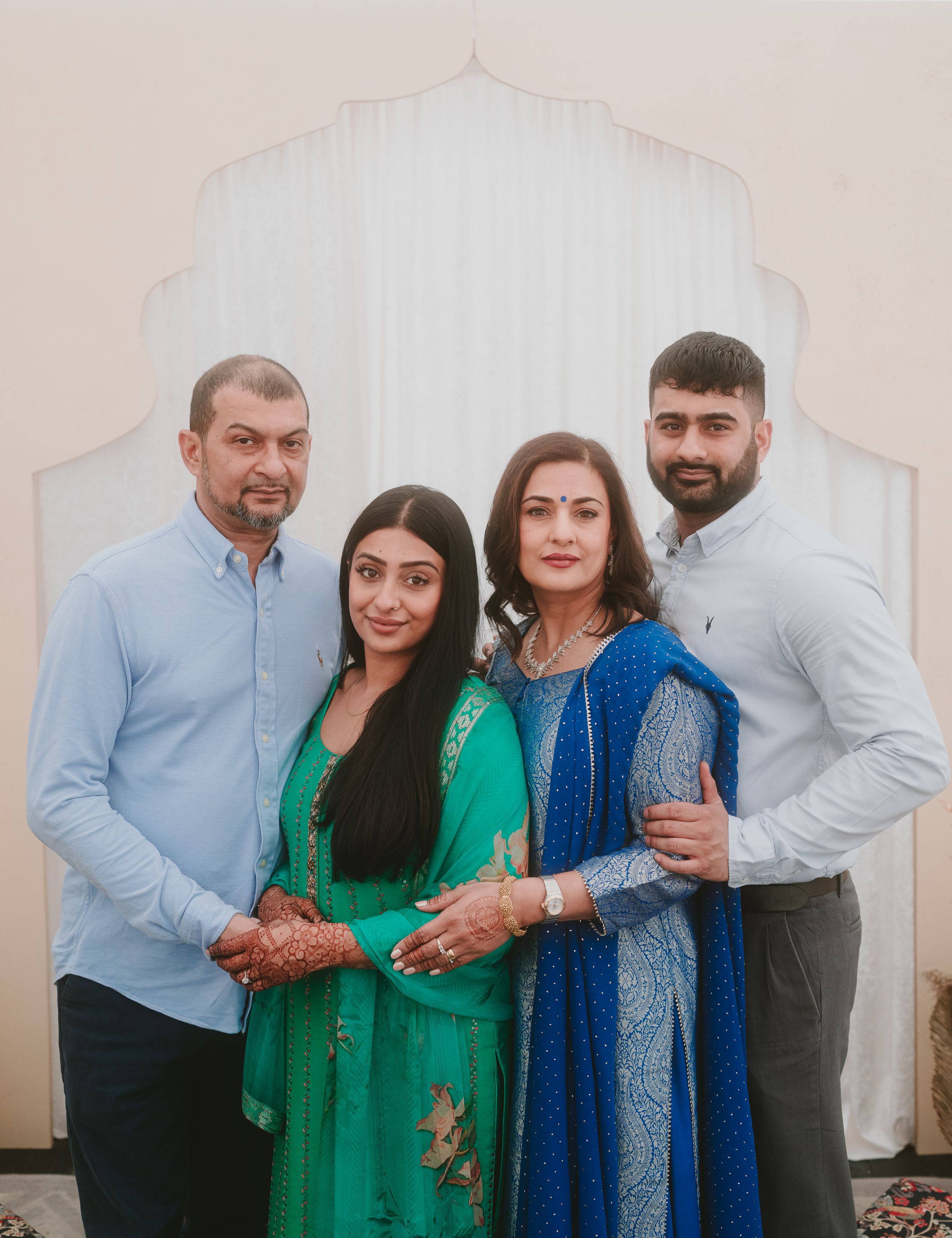 A family of four in traditional South Asian attire poses together against a white backdrop.