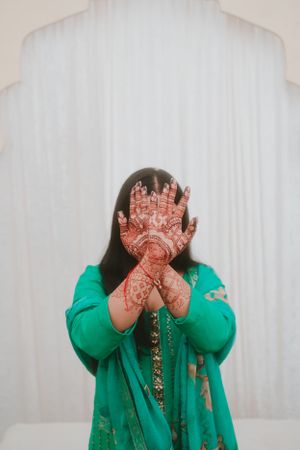 Playful pose with intricate henna-decorated hands covering face against white background.