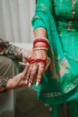 Traditional red bangles being placed on henna-adorned hands wearing a green outfit.