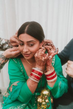Person wearing green outfit and red bangles adjusts jewelry with henna-decorated hands.