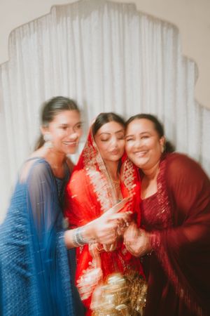 Three women in traditional South Asian attire share a joyful moment against a white backdrop.