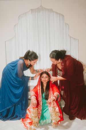 Traditional wedding preparation scene with three people in colorful Indian attire against white backdrop.