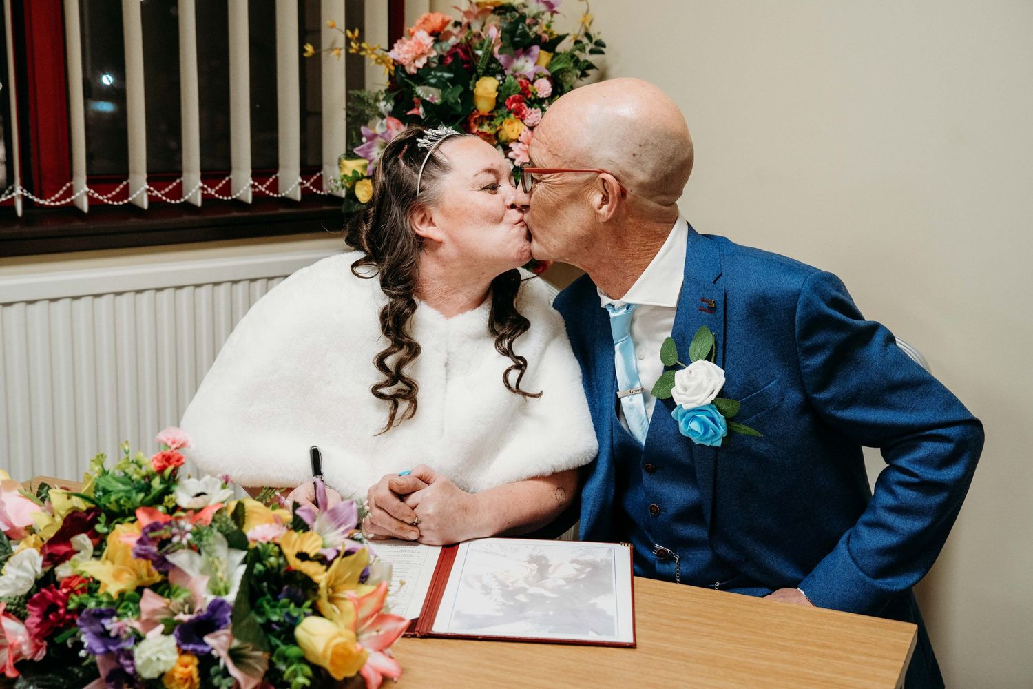 Wedding couple shares intimate moment while signing marriage documents surrounded by floral decor.