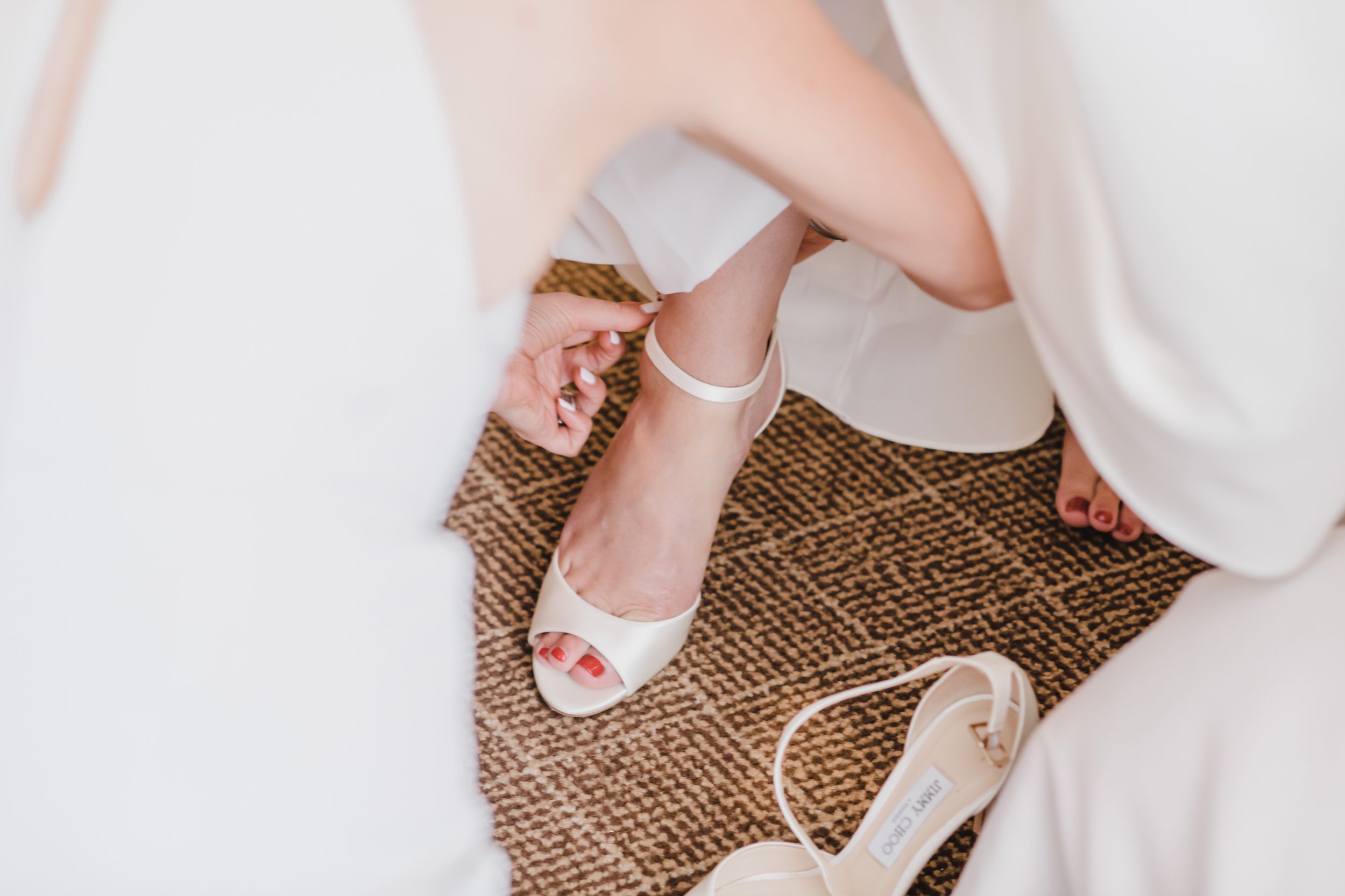 Elegant white wedding shoes with painted pink toenails visible on a textured brown surface.