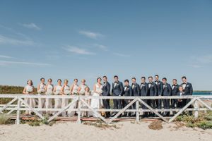 Large wedding party poses on beach boardwalk with bridesmaids in white and groomsmen in navy.