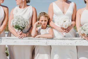 Wedding party members hold white bouquets while standing together on a sunny beach.