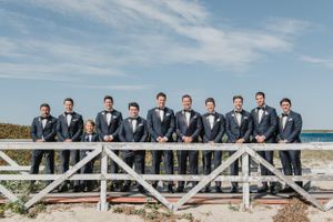 A row of groomsmen in matching grey suits pose on a wooden boardwalk by the ocean.