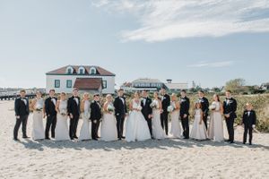 A wedding party poses in a line on the beach with elegant attire and bouquets.